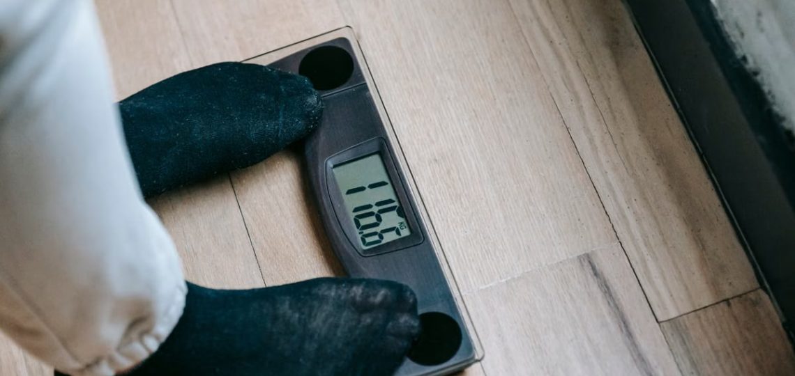 From above of unrecognizable person in socks standing on electronic weighing scales while checking weight on parquet during weight loss